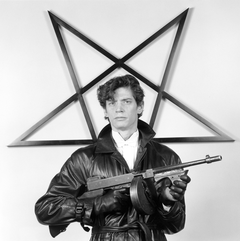 Robert Mapplethorpe wearing a leather jacket and gloves and holding a gun in front of a star sculpture.