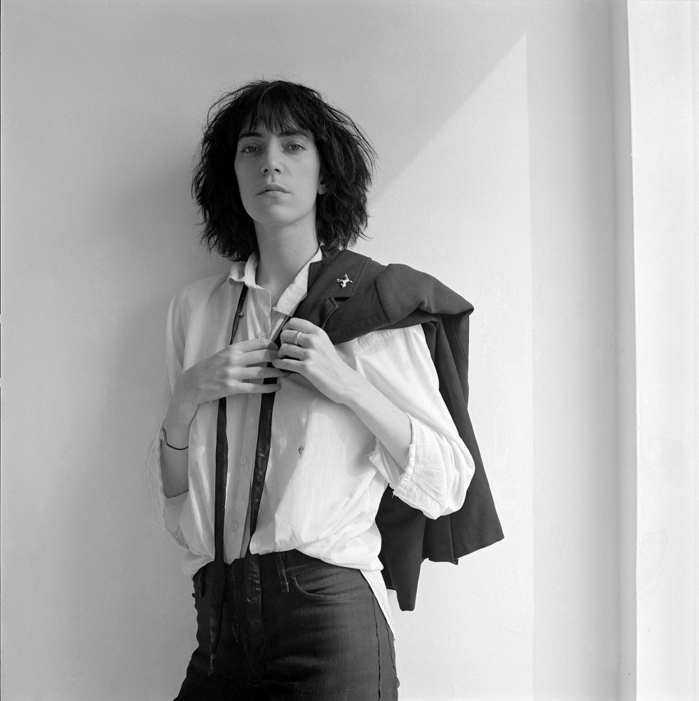 Patti Smith facing camera, suit jacket draped over shoulder, triangular patch of light behind her.