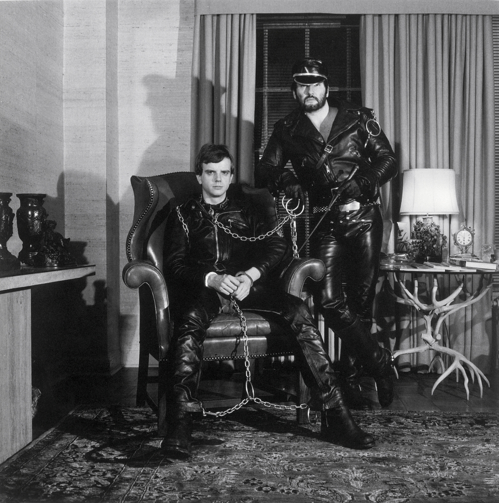 Lyle Heeter and Brian Ridley in full leather and a harness, seated in their living room.