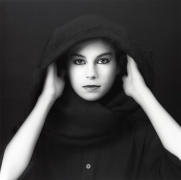 Close-up of a woman in heavy black eyeliner and a black hooded cape with her hands to her ears staring at the viewer.