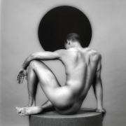 Felix Brown seated on pedestal, nude, facing away from camera towards black sphere.