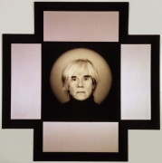 A portrait of the artist Andy Warhol from the neck up in a black turtleneck looking at the viewer. The black frame with white mat around the image forms a cross shape.
