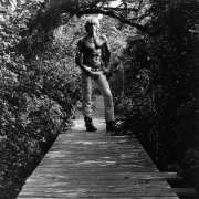 Shirtless white male with hands on hips, standing on boardwalk surrounded by foliage.