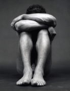 Black male nude sitting down, hands covering face and feet together.