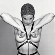 Close-up of woman with exposed breasts, wearing a leather harness and chainmail helmet clenching her fists over her nipples and looking downward.