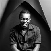 Man wearing a black collared shirt laughing with his eyes closed in front of a dark geometric background.