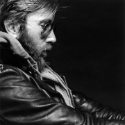 Close-up profile view of a man with a beard wearing sunglasses and a leather jacket over a zip-up sweatshirt.