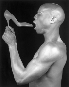 Ken Moody in profile, holding a high heeled shoe close to his mouth.