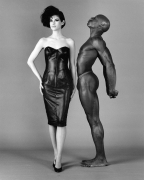A woman in a leather cocktail dress and high heels facing the camera. An almost nude man stands next to her facing her and looking upward.