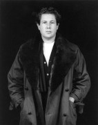 The artist Julian Schnabel from the hips up facing and looking straight at the viewer. His hands are in the pockets of his trench coat with dark shearling lapels.