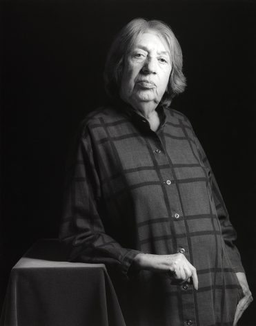 The artist Lee Krasner from the hips up. She wears a dark checked collared shirt, leans against a small table, and looks into the camera.