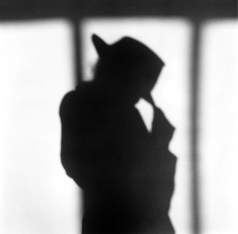 Shadow silhouette of Lisa Lyon in a trench coat and hat against a white wall.