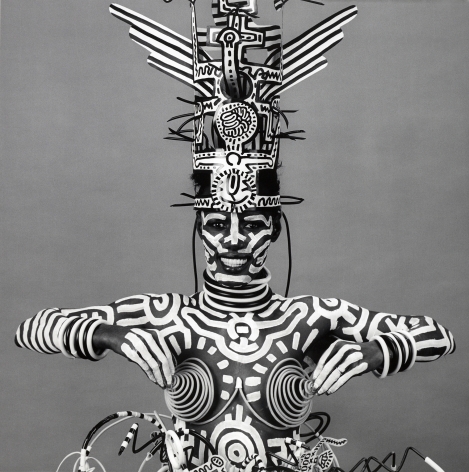 Artist and musician Grace Jones from the waist up looking and smiling into the camera. She is wearing a pointy wire bra and body paint and a hat by Keith Haring and holds the nipples of the bra.