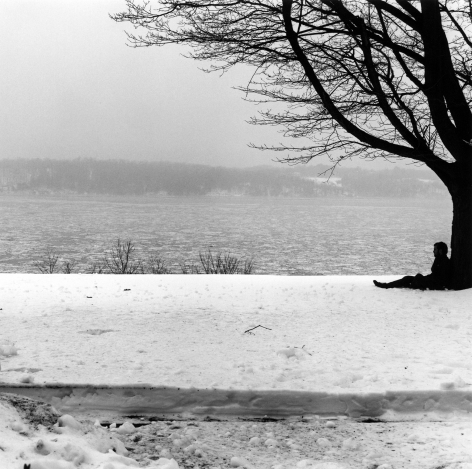 Person in profile sitting against leafless tree on snow covered ground.