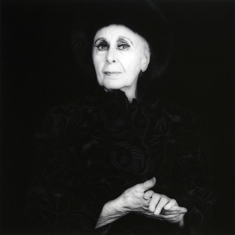 Artist Louise Nevelson from the waist up looking into the camera with her hands clasped in front of her. She wears a black fur hat and a black jacket embroidered with black flowers.