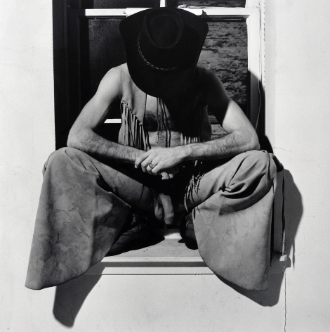 Man crouching in window, wearing cowboy hat, pants, and and vest looking down with penis visible.