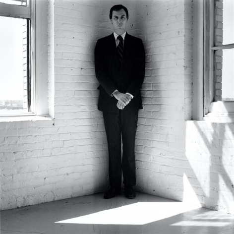 Robert Wilson wearing a suit standing in the corner of a white room.