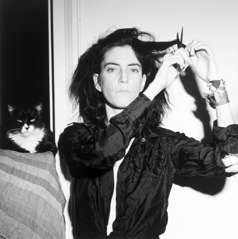 Patti Smith facing camera, cutting her hair, with cat in background.