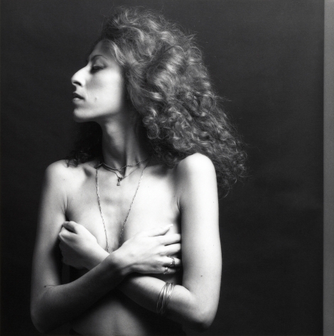 Portrait of a topless woman with curly hair and silver jewelry turning her face in profile and crossing her arms to cover her breasts.