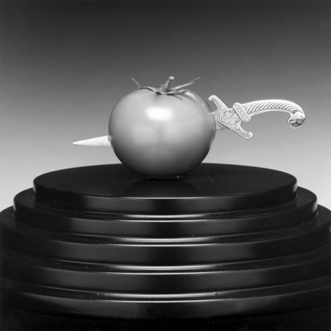 A tomato on a leveled round black stand with a silver dagger pieced though its center.