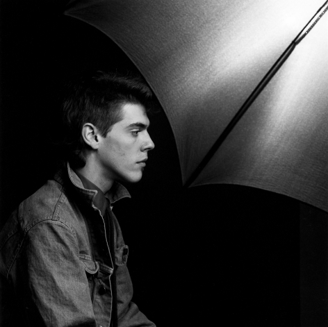 Profile view of young man wearing a denim jacket sitting in front of the visible reflecting umbrella from the studio set-up.