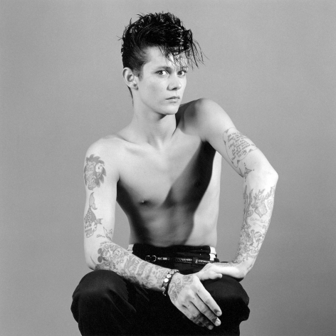 Seated shirtless person with tattoos and gelled hair.