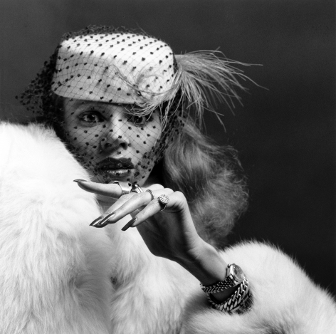 Woman from the shoulders up with a surprised expression wearing a birdcage hat and white fur coat extending her hand up at the center of the image showing her jewelry.