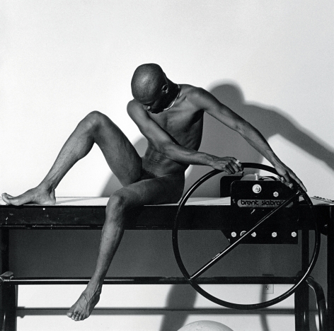Nude black man sitting on a clay slab roller and looking downward.
