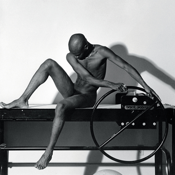 Nude black man sitting with hands on the wheel of a slab roller and looking downward. 
