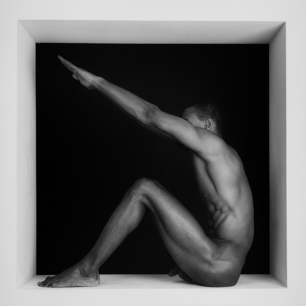 Thomas naked, sitting in a square with arms reaching up..