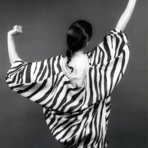 Lisa Lyon in zebra- patterned dress and pearls with hands up, facing away from camera