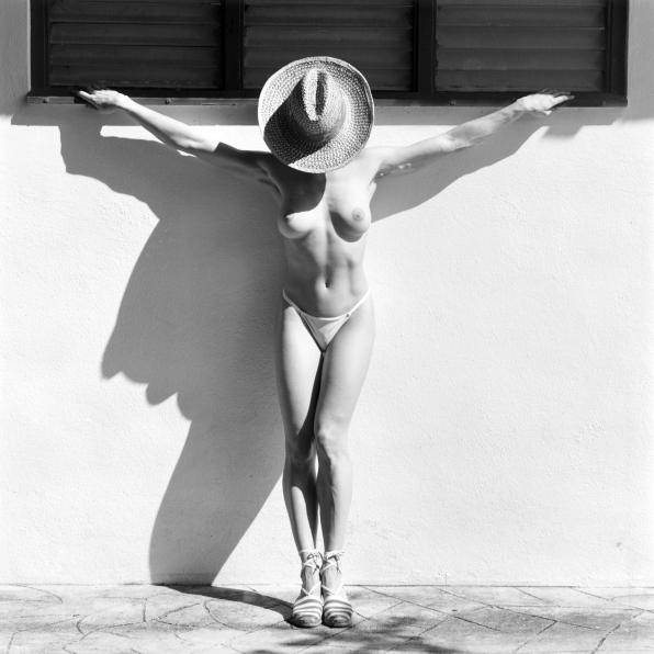 Lisa Lyon standing against a sunny wall with arms outstretched and head down, wearing a bikini bottom, hat,  and espadrilles.