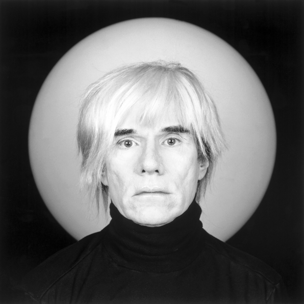 Portrait of Andy Warhol posed in front of a white circle against black background.