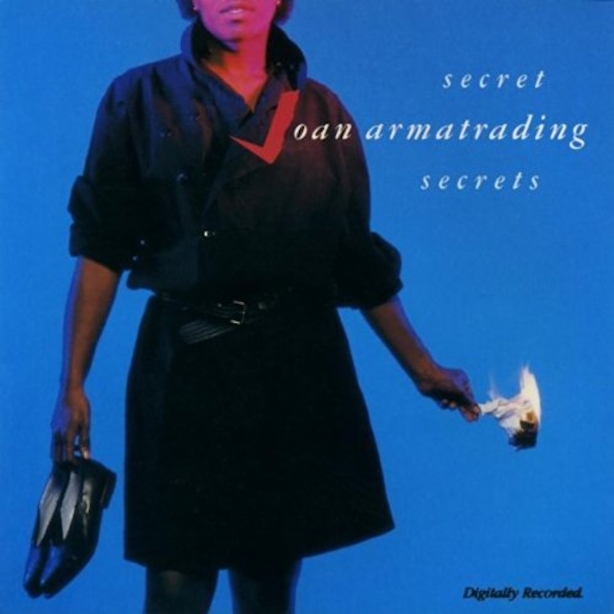 Album cover Joan Armatrading Secret Secrets, Joand in a black dress standing against a blue background with her shoes in her hand.